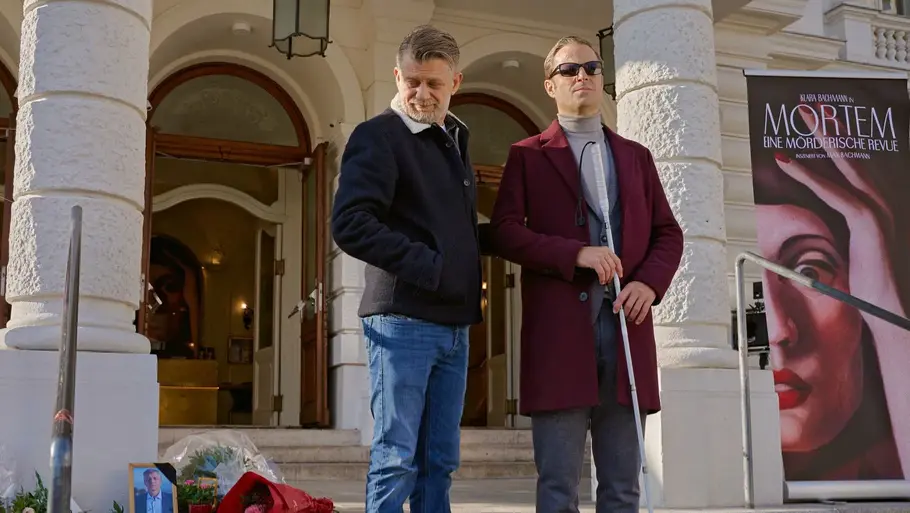 „Blind ermittelt: Tod an der Donau“ heute in der ARD Alexander Haller und Niko Falk stehen vor einem Theatergebäude; Haller hält einen Blindenstock, daneben sind Blumen und ein Filmplakat zu sehen.