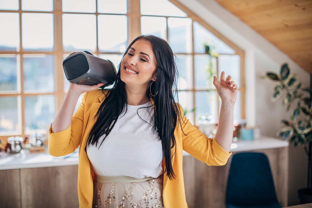 Stiftung Warentest hat ihr Urteil gesprochen: Die besten Bluetooth-Lautsprecher für dich! Eine Frau mit gelber Bluse hält lachend eine Bluetooth-Box über ihre Schulter und bewegt sich dazu rhythmisch.