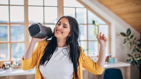 Eine Frau mit gelber Bluse hält lachend eine Bluetooth-Box über ihre Schulter und bewegt sich dazu rhythmisch. - Foto: iStock/ South_agency