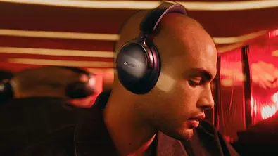 Eine Person mit Bose Over-Ear-Kopfhörern blickt nach unten, während warmes rotes Licht den Innenraum eines Fahrzeugs erhellt. Die Szene vermittelt eine ruhige, abgeschirmte Atmosphäre unterwegs. - Foto: Amazon/PR Eine Person mit Bose Over-Ear-Kopfhörern blickt nach unten, während warmes rotes Licht den Innenraum eines Fahrzeugs erhellt. Die Szene vermittelt eine ruhige, abgeschirmte Atmosphäre unterwegs. - Foto: Amazon/PR