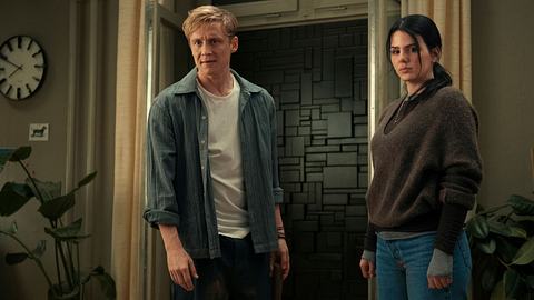 Man sieht Matthias schweighöfer und Ruby O. Fee in einer Wohnung stehen. Vor ihrer Tür steht eine dunkle schwarze Mauer aus Steinen, darüber hinaus sieht am linken und rechten Rand zwei Pflanzen und oben links sieht man eine Uhr. Schweighöfer trägt ein blaues Hemd, eine schwarze Hose und ein weißes T-Shirt, während Fee einen braunen Pulli, sowie eine blaue Jeans aufträgt. - Foto: Netflix
