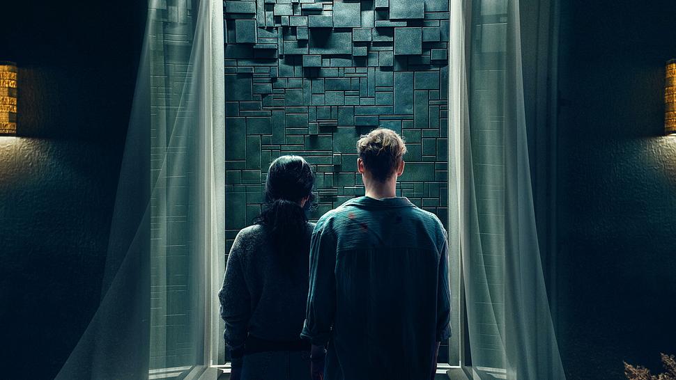 Olivia (links) und Tim stehen vor einer grauen Wand, die von zwei Vorhängen umhüllt wird. - Foto: Netflix
