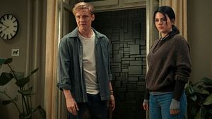 Matthias Schweighöfer und Ruby O. Fee stehen in einer Wohnung. Oben links ist eine Uhr, unten links eine Topfpflanze. Schweighöfer trägt ein blaues Hemd, weißes Shirt und eine schwarze Hose. Fee trägt einen braunen Hoodie und eine blaue Jeans. im Hintergrund sieht man eine graue Mauer, die die Tür versperrt. - Foto: Netflix