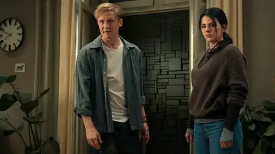 Matthias Schweighöfer und Ruby O. Fee stehen in einer Wohnung. Oben links ist eine Uhr, unten links eine Topfpflanze. Schweighöfer trägt ein blaues Hemd, weißes Shirt und eine schwarze Hose. Fee trägt einen braunen Hoodie und eine blaue Jeans. im Hintergrund sieht man eine graue Mauer, die die Tür versperrt. - Foto: Netflix