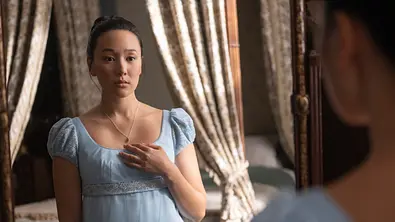 Yerin Ha trägt als Sophie Baek in der 4. Staffel von „Bridgerton“ ein hellblaues, elegantes Kleid, eine schlichte Kette und schaut ihr Spiegelbild an. - Foto: Liam Daniel/Netflix © 2025