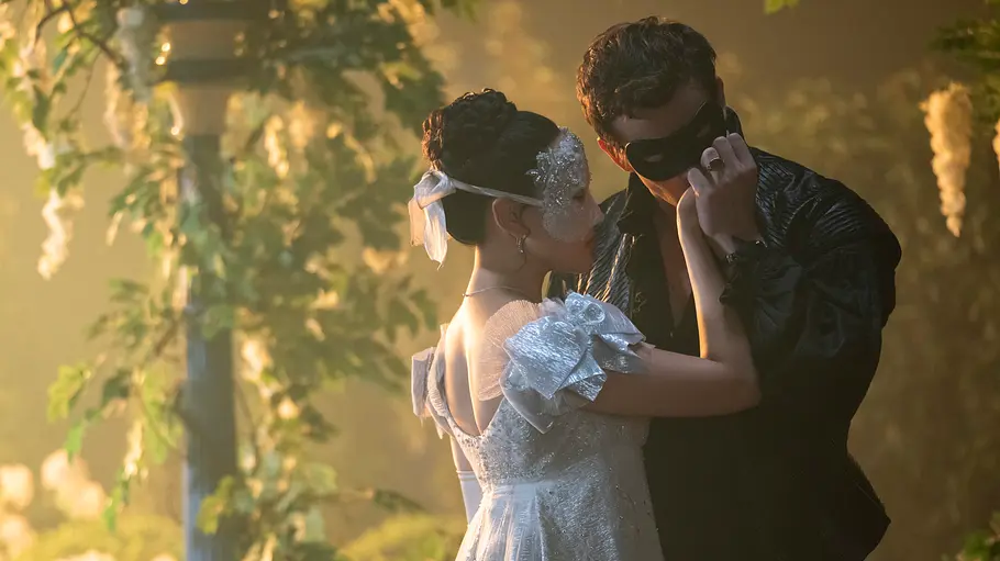 Sophie und Benedict Eine junge Frau in einem glitzernden, weißen Ballkleid und mit filigraner Halbmaske steht einem jungen Mann in dunkler Kleidung und schwarzer Maske gegenüber. Die beiden sind einander ganz nah und halten sich zärtlich an den Händen, im Hintergrund ranken grüne Pflanzen und weiße Blüten an einem Pavillon. Die Szene wirkt geheimnisvoll und romantisch.