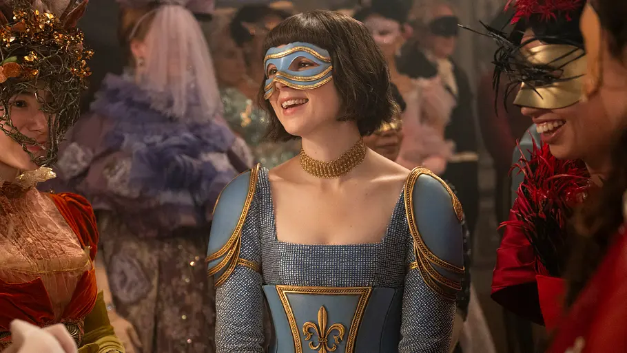 Eloise in „Bridgerton“ Mehrere Personen in aufwendigen historischen Kostümen und fantasievollen Masken stehen zusammen auf einem Ball und unterhalten sich. Die zentrale Figur trägt ein blau-goldenes Kostüm mit Wappen und eine passende Gesichtsmaske, während die anderen ebenfalls aufwendig gekleidet und maskiert sind. Die Stimmung ist fröhlich und ausgelassen.