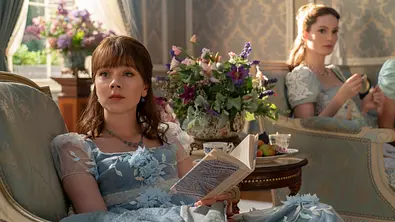 Zwei junge Frauen in eleganten, hellblauen, historisch inspirierten Kleidern sitzen in einem prächtig dekorierten Salon auf prunkvollen Sesseln. Die Frau im Vordergrund hält ein aufgeschlagenes Buch und blickt gedankenverloren nach vorne. Im Hintergrund sitzt die zweite Frau, die eine Tasse hält. Zwischen ihnen steht ein Tisch mit einem Blumenstrauß und einer Schale mit Obst. Die Atmosphäre wirkt ruhig und stilvoll. - Foto: Netflix Zwei junge Frauen in eleganten, hellblauen, historisch inspirierten Kleidern sitzen in einem prächtig dekorierten Salon auf prunkvollen Sesseln. Die Frau im Vordergrund hält ein aufgeschlagenes Buch und blickt gedankenverloren nach vorne. Im Hintergrund sitzt die zweite Frau, die eine Tasse hält. Zwischen ihnen steht ein Tisch mit einem Blumenstrauß und einer Schale mit Obst. Die Atmosphäre wirkt ruhig und stilvoll. - Foto: Netflix