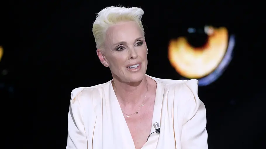 Brigitte Nielsen trägt kurzes blondes Haar und ein weißes Oberteil mit Ansteckmikrofon vor dunklem Hintergrund.