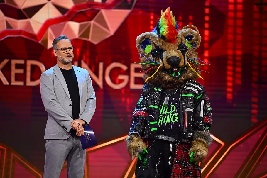 Bunt, punkig und bewusst auffällig: Wild Thing präsentiert sich als rebellischer Neon-Hingucker auf der „Masked Singer“-Bühne. Matthias Opdenhövel steht im grauen Anzug neben Wild Thing im pelzigen Punkkostüm mit buntem Irokesenschnitt auf einer roten Bühne.