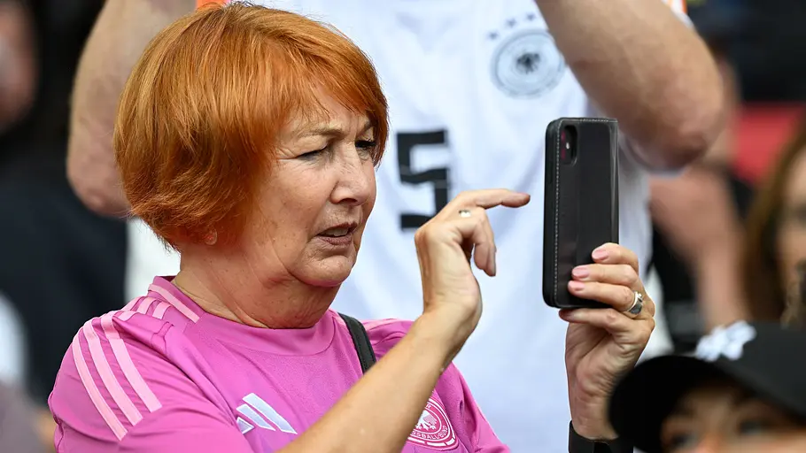 Brugi Nagelsmann Eine Fußballfanin im pinken Trikot der deutschen Nationalmannschaft hält auf der Tribüne ihr Smartphone hoch und macht ein Foto oder Video. Mit konzentriertem Blick richtet sie die Kamera aus, während um sie herum weitere Zuschauer das Spielgeschehen im Stadion verfolgen.