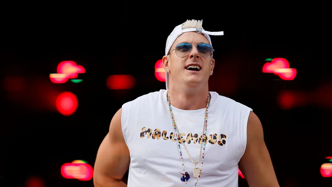 Reality-TV-Star Calvin Kleinen steht auf einer Bühne vor dunklem Hintergrund mit roten Lichteffekten. Er trägt ein weißes ärmelloses Shirt mit Aufdruck, eine rückwärts getragene weiße Basecap, eine Sonnenbrille sowie mehrere Ketten und blickt während seines Auftritts ins Publikum. - Foto: IMAGO / Panama Pictures