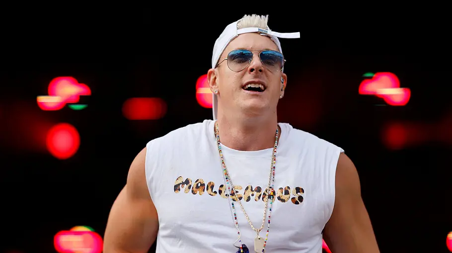 Calvin Kleinen Reality-TV-Star Calvin Kleinen steht auf einer Bühne vor dunklem Hintergrund mit roten Lichteffekten. Er trägt ein weißes ärmelloses Shirt mit Aufdruck, eine rückwärts getragene weiße Basecap, eine Sonnenbrille sowie mehrere Ketten und blickt während seines Auftritts ins Publikum.