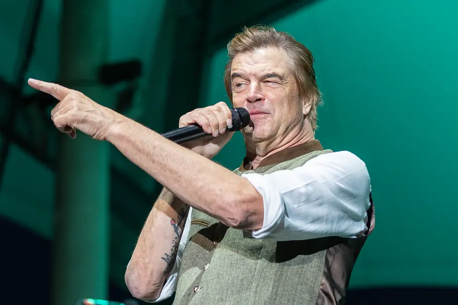 Campino Tote Hosen-Frontsänger Campino steht auf einer Bühne, hält ein Mikrofon in der rechten Hand und zeigt mit ausgestrecktem linken Arm zur Seite.