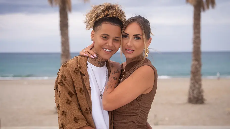 Carina Nagel und Chika Ojiudo-Ambrose sind das erste queere Paar der Sendung. Carin und Chika in brauner Kleidung umarmen sich vor einem Sandstrand mit Palmen und Meer unter bewölktem Himmel.