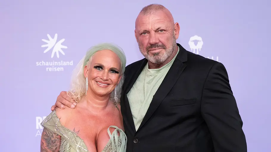 Caro und Andreas Robens Caro Robens im verzierten hellgrünen Kleid und Andreas Robens im schwarzen Sakko posieren eng beieinander vor einer Logowand.