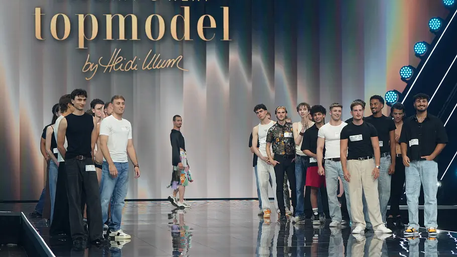 Casting der Männer bei GNTM 2026 Eine Gruppe männlicher Kandidaten steht auf dem Catwalk bei „Germany's Next Topmodel“ vor dem großen „topmodel by Heidi Klum“-Schriftzug. Im Hintergrund laufen weitere Teilnehmer auf dem Laufsteg, die Szene ist von Scheinwerfern und Studio-Lichtern beleuchtet.