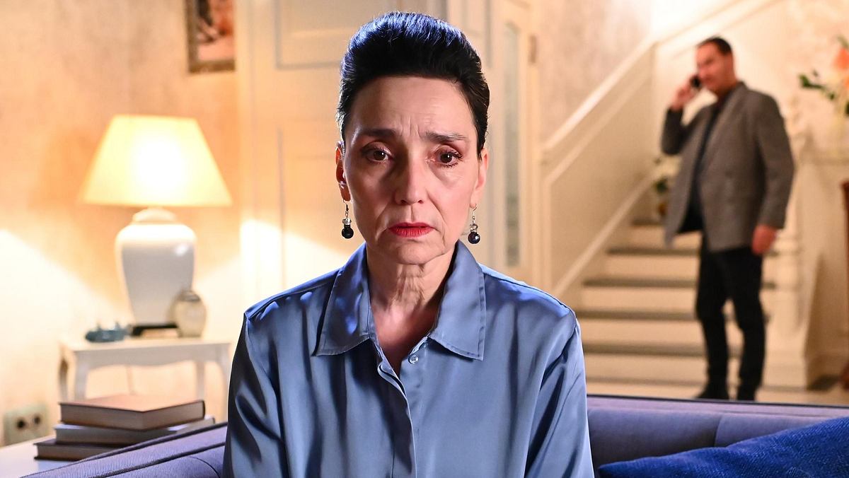 Simone sitzt auf der Couch in ihrer Villa und schaut traurig in Richtung Kamera, im Hintergrund telefoniert Richard und schaut in ihre Richtung bei Alles was zählt