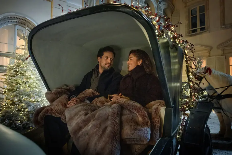Chamapgne Problems Netflix Tom Wozniczka als Henri Cassell und Minka Kelly als Sydney Price sitzen in einer Art Kutsche mit einer dicken Decke und weihnachtlicher Dekoration. Sie schauen sich lächelnd an.