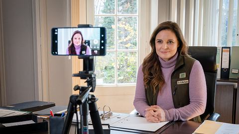 Dr. Mertens sitzt an ihrem Schreibtisch, mit einem Handy auf einem Stativ nimmt sie sich auf. - Foto: © ARD/Saxonia Media/Steffen Junghans