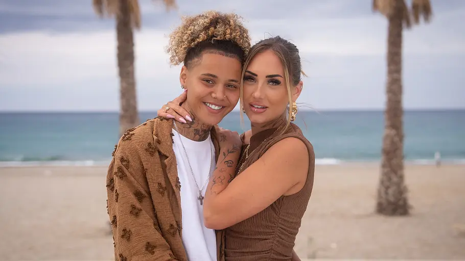 Chika und Carina Chika und Carina stehen eng umschlungen vor einem Strand mit Palmen und Meer und schauen in die Kamera.