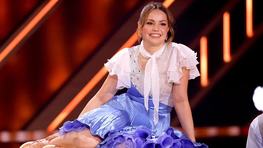 Christina Hänni Not-OP. Christina Hänni im blau-weißen Rüschenkleid und goldfarbenen Tanzschuhen sitzt vor orangefarbenen Lichtstreifen.