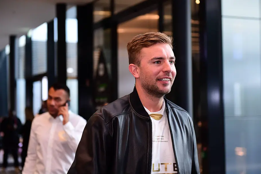 Christoph Kramer Christoph Kramer steht in einem Flur. Im Hintergrund telefoniert ein Mann im weißen Hemd.