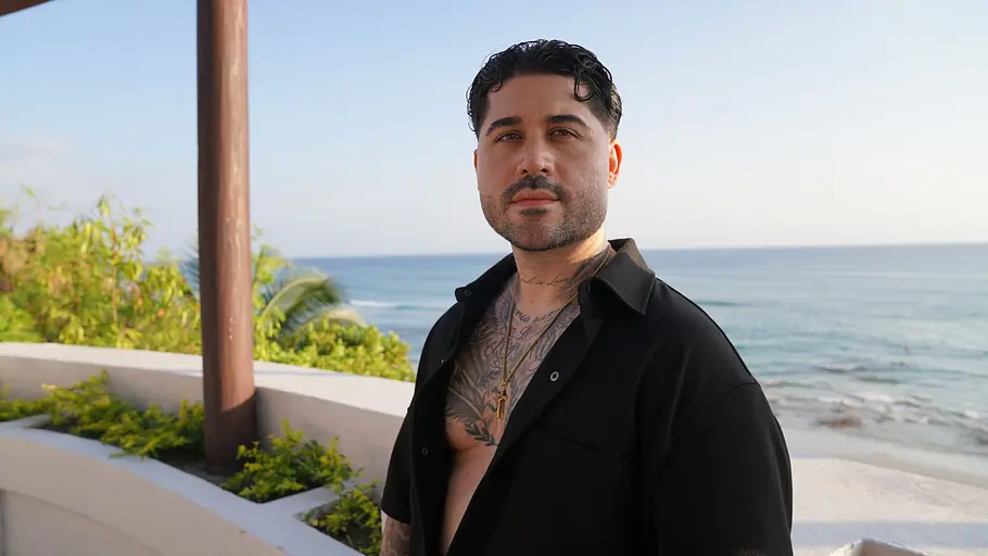 Christos steht in einem offenen, schwarzen Hemd vor Urlaubs-Kulisse.