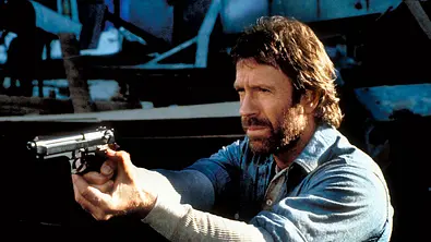 Chuck Norris steht mit einer Waffe da und zielt konzentriert in die Ferne. - Foto:  IMAGO / Everett Collection