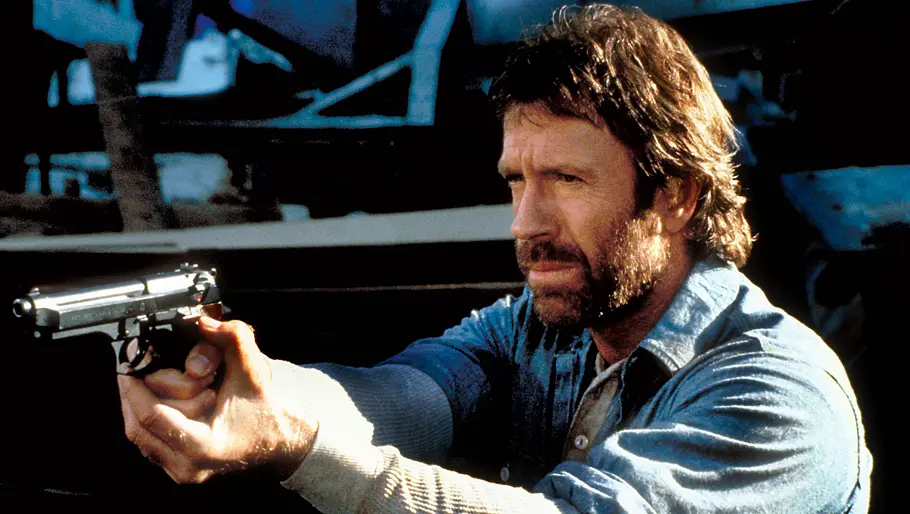 Chuck Norris steht mit einer Waffe da und zielt konzentriert in die Ferne.