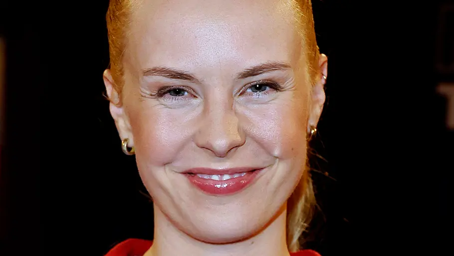 Eine lächelnde Frau mit zurückgebundenen blonden Haaren trägt eine rote Strickjacke vor dunklem Hintergrund.