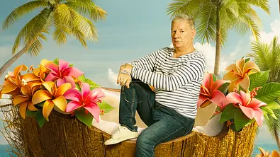 Ein Mann sitzt auf einer halbierten Kokosnuss an einem tropischen Strand, umgeben von Palmen und bunten Blumen. Im Hintergrund ist das Meer zu sehen, darüber fliegt ein Papagei. - Foto: Joyn