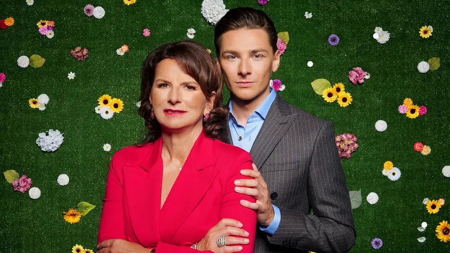 Claudia Obert und Max Suhr Claudia Obert im roten Blazer und Max Suhr im grauen Nadelstreifenanzug posieren vor einer grünen Wand mit bunten Blumen.