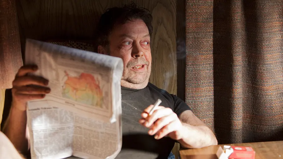 Criminal Minds Ein Mann sitzt mit einer Zigarette und einer Zeitung in einem Wohnwagen und blickt nachdenklich aus dem Fenster. Auf dem Tisch liegen eine Zigarettenschachtel und ein Aschenbecher – Szene aus „Criminal Minds“.