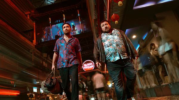 Man sieht Frederick Lau (links) und Christoph Krutzler, die durch eine neon-beleuchtete Gasse in Thailand gehen. Lau trägt eine Tasche in seiner Hand und ein blau-rot gemustertes Hemd. Krutzler trägt ein Hawaii-Hemd und darüber eine Jacke. - Foto: Sasidis Sasisakulporn/Netflix