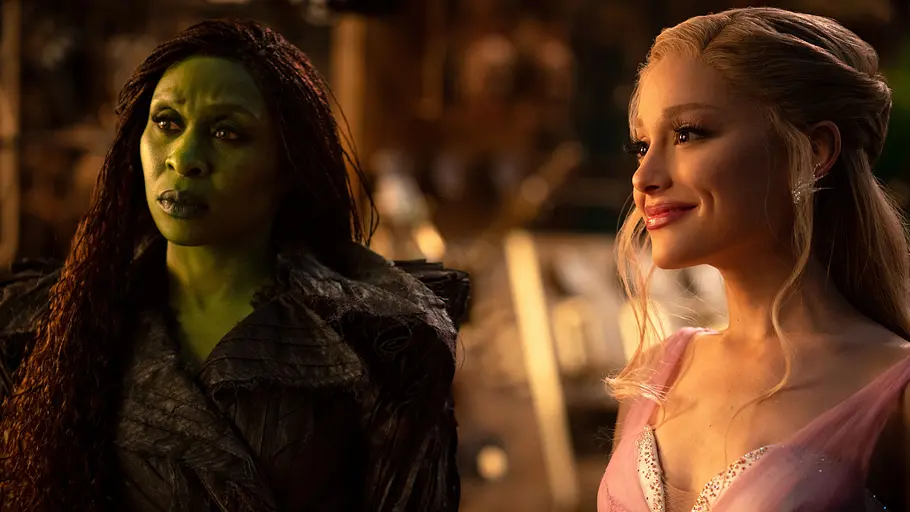 Cynthia Erivo als Elphaba und Ariana Grande als Glinda in Wicked Cynthia Erivo als Elphaba schaut skeptisch. Sie hat grüne Haut und trägt ein schwarzes Gewand. Ariana Grande als Glinda lächelt freundlich, strahlt und trägt ein rosa Kleid.