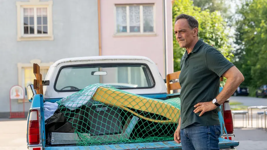 Mike steht mit einer Hand an der Hüfte neben einem blauen Pick-up, dessen beladene Ladefläche mit einem grünen Netz gesichert ist.