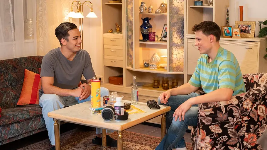 Leon und Hacki sitzen sich lächelnd auf Sofa und Sessel gegenüber. Auf dem Wohnzimmertisch liegen Snacks und Kopfhörer.