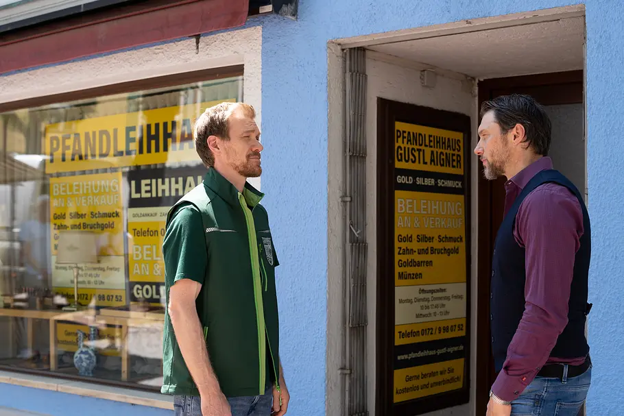 Charly und Aigner stehen sich vor einem Pfandleihhaus mit blauer Fassade und gelben Schildern gegenüber.