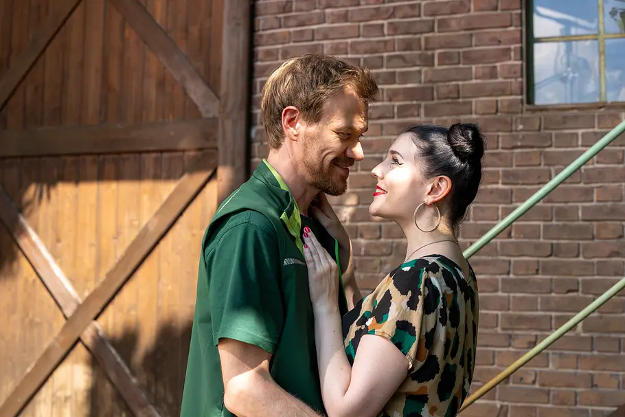 Charly und Tina stehen eng beieinander vor einer Backsteinwand und einem Holztor und schauen sich lächelnd an.