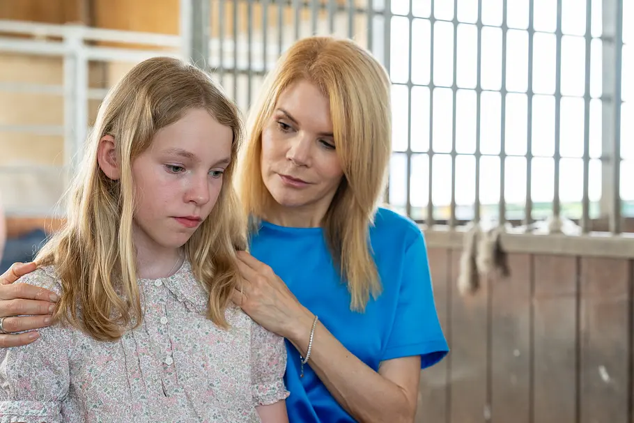 Uschi im blauen Oberteil legt Franzi die Hände auf die Schultern. Franzi trägt eine geblümte Bluse und blickt nach unten.