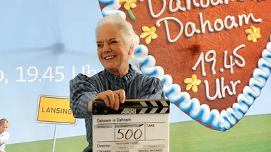 Ursula Erber hält eine Filmklappe am Set von „Dahoam is Dahoam“ vor einem Hintergrund mit Serienlogo und Sendetermin. - Foto: BR/Marco Orlando Pichler