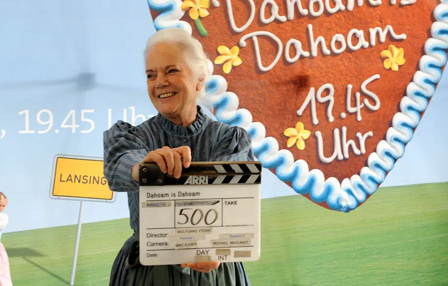 Ursula Erber hält eine Filmklappe am Set von „Dahoam is Dahoam“ vor einem Hintergrund mit Serienlogo und Sendetermin.