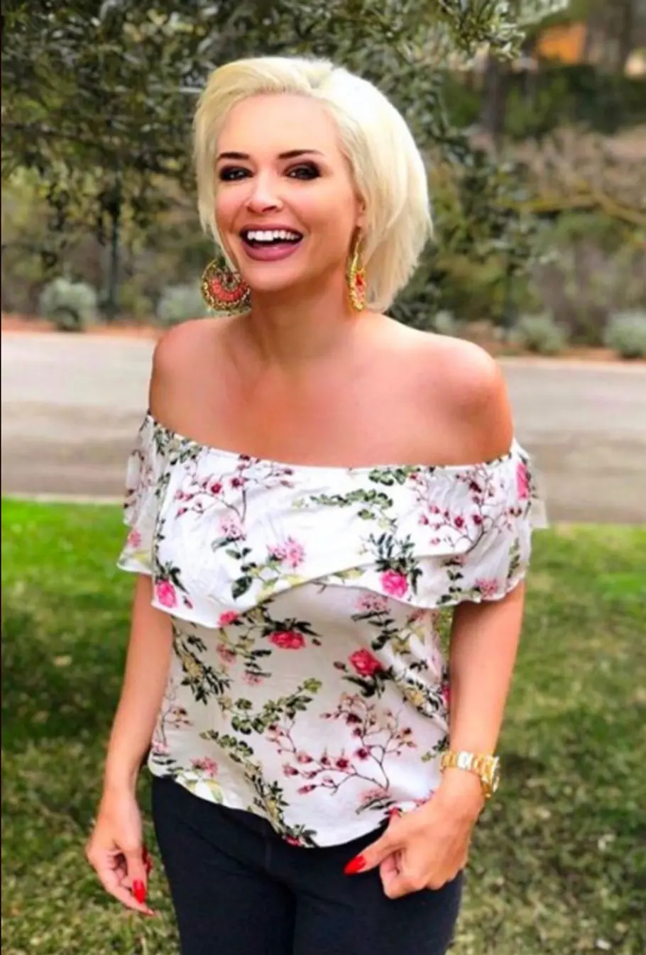 Daniela Katzenberger ohne Extensions Daniela Katzenberger lächelt im Freien mit blondem Bob, großen Ohrringen und einer weißen, schulterfreien Bluse mit Blumenmuster. Ohne Extensions sieht die Katze ganz anders aus.