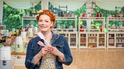 Eine rothaarige Frau mit geflochtenem Haar steht in einer hellen Backstube und hält einen Schneebesen sowie zwei rosa Teigschaber in den Händen. Sie trägt ein geblümtes Kleid mit Jeansjacke, im Hintergrund sind Regale mit Backzutaten und Küchenutensilien zu sehen. - Foto: SAT.1 / Claudius Pflug