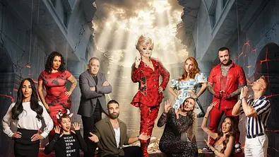 Gruppenbild eines Reality-TV-Casts vor einer dramatischen Kulisse: In einem beschädigten Gebäude mit eingestürzter Decke steht eine Person in einem auffälligen roten Outfit zentral auf einer Treppe und wird von Lichtstrahlen von oben beleuchtet. Um sie herum posieren mehrere weitere Personen in unterschiedlich auffälligen Outfits, einige mit betenden oder überraschten Gesten. Die Szenerie wirkt theatralisch und apokalyptisch, mit Rissen in den Wänden und rot glühenden Effekten im Hintergrund. - Foto: Joyn/Nikolai Milatovic