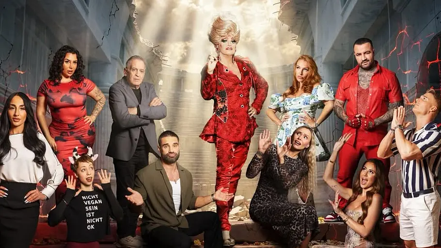 Promi Büßen Gruppenbild eines Reality-TV-Casts vor einer dramatischen Kulisse: In einem beschädigten Gebäude mit eingestürzter Decke steht eine Person in einem auffälligen roten Outfit zentral auf einer Treppe und wird von Lichtstrahlen von oben beleuchtet. Um sie herum posieren mehrere weitere Personen in unterschiedlich auffälligen Outfits, einige mit betenden oder überraschten Gesten. Die Szenerie wirkt theatralisch und apokalyptisch, mit Rissen in den Wänden und rot glühenden Effekten im Hintergrund.