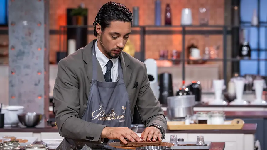 René Casselly arbeitet konzentriert in einer TV-Backshow in der Studioküche und bereitet einen Kuchen auf einer Tortenplatte zu.