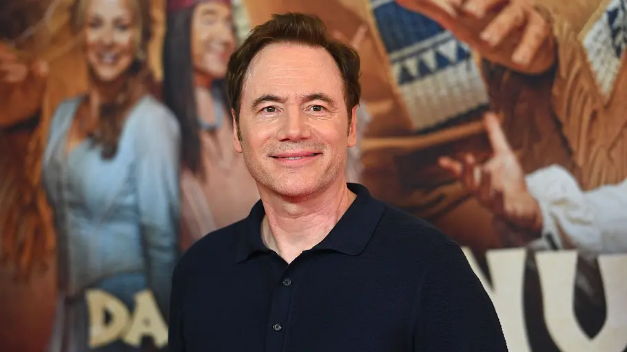 Dass der große Erfolg von „Das Kanu des Manitu“ kein glücklicher Zufall war, hat Michael „Bully“ Herbig mittlerweile mehrfach bewiesen. Michael „Bully“ Herbig lächelt in einem dunkelblauen Poloshirt vor einem Filmplakat mit mehreren Figuren.