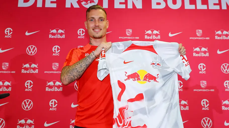 David Raum David Raum hält vor einer roten Fotowand ein Trikot des RB Leipzig hoch.